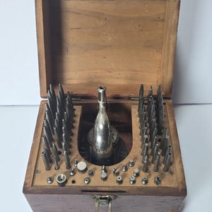 May include: A wooden storage box containing a collection of metal watchmaker tools. The set includes numerous pointed steel punches and a central silver-toned metal staking tool. The box features a hinged lid and a metal latch on the front.