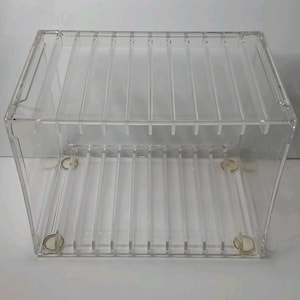 May include: A clear acrylic storage organizer featuring a rectangular frame with multiple vertical dividers creating individual slots. The structure is transparent, showcasing a grid-like interior design with four small circular feet at the base.
