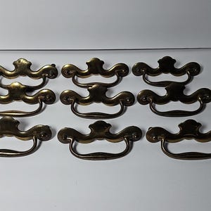 May include: A set of nine brass drawer pulls arranged in three rows of three. Each pull features a decorative backplate with a curved, ornate top edge and a matching swinging bail handle attached to two mounting points on the sides.