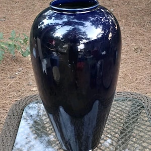 May include: A large, glossy, deep navy blue ceramic vase with a smooth, reflective surface. The vase features a rounded, bulbous body that tapers toward a narrow, circular opening at the top, showcasing a solid, dark, and uniform color finish.