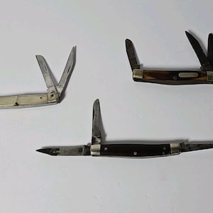 May include: Three folding pocket knives with metallic blades and handles in varying shades of cream, brown, and dark wood. Each knife features multiple blades extended at different angles, with metallic bolsters and rivets visible on the handles.