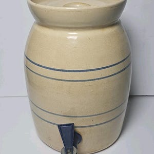 Op de afbeelding: Een cilindervormige beige stenen kruik met een bijpassend deksel en een donkerblauwe kunststof kraan aan de onderkant. De buitenkant is voorzien van twee sets dunne, horizontale blauwe strepen die rondom het lichaam van de kruik zijn aangebracht.