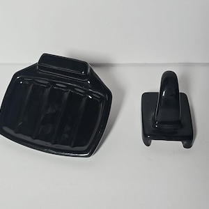 Könnte beinhalten: Ein schwarzes Keramik-Seifenschalen- und Seifenhalter-Set. Die Seifenschale hat eine rechteckige Form mit abgeschrägtem Design und Rillen. Der Seifenhalter hat einen rechteckigen Sockel mit einem gebogenen Griff.