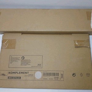 May include: Two brown cardboard boxes, one rectangular and one square, are stacked on top of each other. The square box has the word "KOMPLEMENT" printed on it, along with measurements of 19.6 x 22.8 inches.