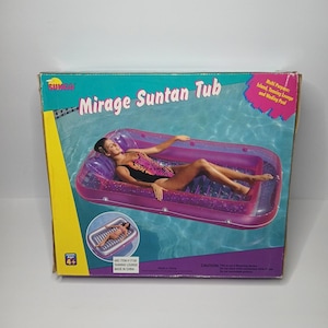 May include: A rectangular cardboard product box for a Sunco Mirage Suntan Tub. The box features a graphic of a purple inflatable pool float on water, with text including Mirage Suntan Tub, Multi Purpose: Island, Tanning Lounge and Wading Pool.