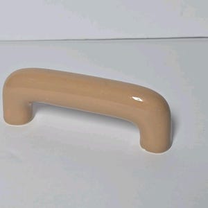 May include: A smooth, light beige ceramic cabinet handle with a rounded, arch-shaped design. The handle features a glossy, reflective finish and two cylindrical mounting points on either end, providing a simple and functional hardware piece.