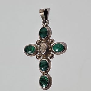 Puede incluir: Colgante plateado en forma de cruz, con piedras ovaladas de malaquita. La cruz tiene un lazo para una cadena. Las piedras de malaquita son de color verde oscuro con patrones arremolinados. Una joya religiosa.