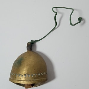 Könnte beinhalten: Eine kleine Messingglocke mit einem grünen Faden. Die Glocke hat ein dekoratives Muster am unteren Rand. Die Glocke hat eine warme Goldfarbe und ein leicht gealtertes Aussehen.