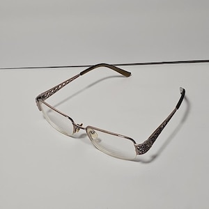 May include: Eyeglasses with a rectangular frame in a light brown hue. The temples feature decorative detailing, and the lenses are clear. The arms are a darker brown colour.