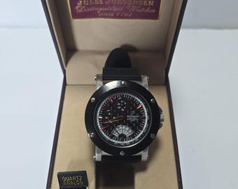 May include: A Jules Jurgensen quartz analog wristwatch with a black bezel, silver-tone case, and black strap, presented in a dark brown hinged box. The watch face features a black textured dial with multiple sub-dials and white markers.