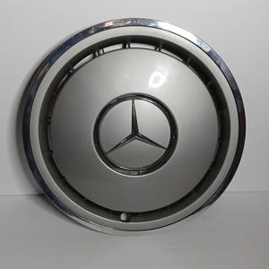 Puede incluir: Tapacubos plateado y cromado con el logotipo de Mercedes-Benz. El tapacubos tiene un diseño circular con un centro plateado y un anillo exterior cromado. El logotipo de Mercedes-Benz es una estrella de tres puntas.