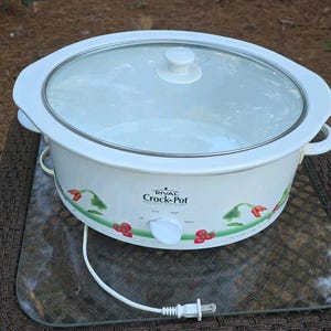 Puede incluir: Una olla de cocción lenta Rival Crock-Pot blanca con tapa de cristal transparente y un mando blanco. El exterior presenta un patrón decorativo de tomates rojos y enredaderas verdes. Un cable de alimentación blanco con enchufe sale de la base.