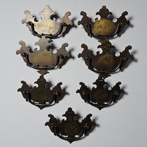 Puede incluir: Un juego de siete tiradores de cajón de latón, cada uno con una placa posterior ornamentada y simétrica con un borde superior curvo y decorativo, y un asa basculante a juego unida a dos pequeños puntos de montaje circulares.