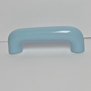 May include: A light blue cabinet handle with a smooth, glossy finish. The handle features a simple, arched bridge shape with two rounded ends that extend downward, providing a clean and uniform appearance for furniture or cabinetry hardware.