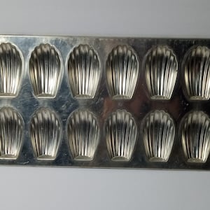 Puede incluir: Un molde rectangular plateado para madeleines con doce moldes en forma de concha. Los moldes tienen un diseño acanalado y la bandeja es de metal brillante. Ideal para hornear madeleines francesas.