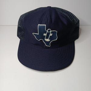 May include: A navy blue trucker cap featuring a mesh back and a solid front panel. Centered on the front is a white-outlined patch shaped like the state of Texas, containing a white graphic of a lowercase letter i with a dot above it.