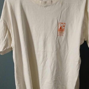 Könnte beinhalten: Cremefarbenes Kurzarm-T-Shirt mit Rundhalsausschnitt. Das Shirt hat ein kleines orange-weißes Logo auf der Vorderseite mit der Aufschrift "PC Magazine Editors' Choice". Das T-Shirt ist aus weichem Material, wahrscheinlich Baumwolle.