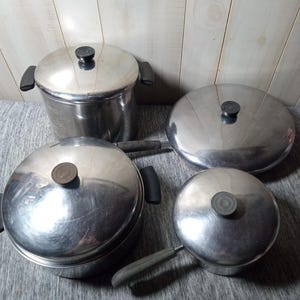 May include: A collection of four stainless steel cooking pots and pans with lids. The set includes a tall pot, two round pots, and a small saucepan. Each piece has a silver finish and black handles or knobs.
