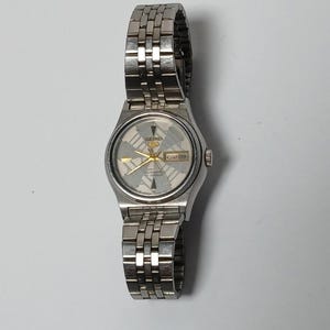 May include: A silver-toned Seiko watch with a patterned face and a metal band. The watch face has gold-colored hands and markers, and a day-date display. The watch is shown on a white background.