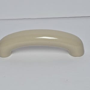 May include: A smooth, arched cabinet handle with a glossy, light beige finish. The hardware features a curved, semi-circular design with rounded ends, providing a clean and simple aesthetic for furniture or cabinetry applications.