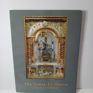 May include: A gray program booklet for The Santa Fe Opera 2004 Festival Season. The cover features a central rectangular illustration of a gold-toned, ornate musical display containing a mandolin, brass instruments, and blue ribbons.