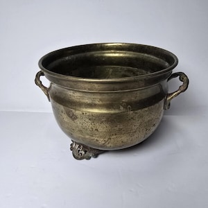 May include: A round brass bowl with a weathered, tarnished patina. The vessel features two small, curved side handles and rests on three ornate, cast metal feet. The interior is deep and smooth, while the exterior surface shows signs of age.