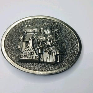 May include: Silver-toned oval belt buckle with a detailed engine relief. The buckle has a textured surface and the text "KHD DEUTZ" is visible. The engine design is highly detailed.