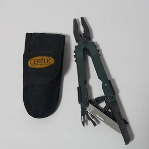 Pode incluir: Uma multi-ferramenta com cabo preto e verde, com alicates, uma faca e outras ferramentas, ao lado de uma bolsa de transporte de nylon preta com o logotipo "GERBER".