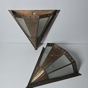 May include: Two triangular wall-mounted sconces crafted from bronze-toned metal and glass panels. Each piece features a central star motif, a fluted base, and a decorative border along the top edge adorned with small cut-out star shapes.