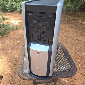 Op de afbeelding: Een verticale computerbehuizing met een zwarte en zilveren buitenkant. Het voorpaneel bevat een dvd-station, een cd-station, twee externe schijfstations, twee USB-poorten en een centrale aan-uitknop op een zilverkleurige voorplaat met een logo.
