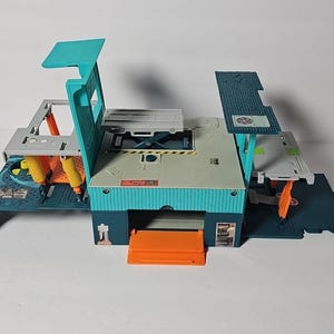 May include: A multi-level plastic toy playset featuring a central teal and gray structure with orange accents. The set includes a fold-out base, a raised platform, a garage-style opening, and various mechanical details like yellow supports and a textured roof.