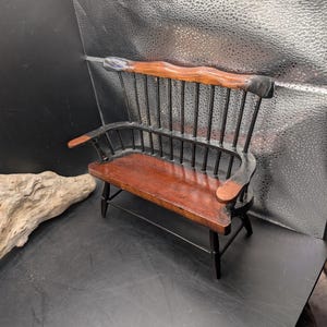 May include: Miniature wooden bench with a curved back and armrests. The seat and backrest are a rich brown, while the frame and spindles are black. The top of the backrest is a lighter brown with blue accents.