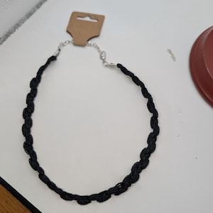 May include: A black lace choker necklace with a silver-colored clasp and a brown tag. The necklace is made of a woven, scalloped lace design. The necklace is displayed on a white surface.