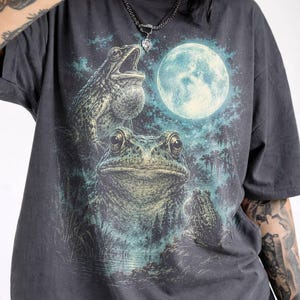 Può includere: T-shirt grigio scuro con un'illustrazione dettagliata di tre rane sotto la luna piena. Il design include una grande luna luminosa, piante acquatiche e uno sfondo nebbioso dai toni blu. Le rane sono in varie pose.