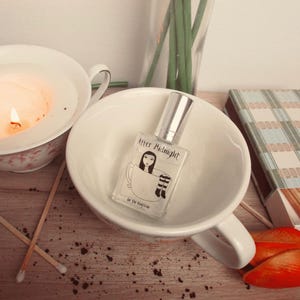 May include: A perfume bottle with a silver cap sits inside a white teacup. The bottle has a label with a drawing of a woman in a teacup and the words "After Midnight". A lit candle and matches are also visible.
