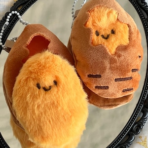 May include: Two plush keychains, one with a fluffy orange creature emerging from a brown egg, and the other with the creature outside the egg. Both have smiling faces and are on a reflective surface.
