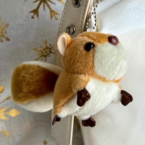May include: A plush, brown and white squirrel keychain with black eyes and a brown nose. The keychain is attached to a cream-colored purse strap with silver hardware. The background features a white fabric with gold snowflake patterns.