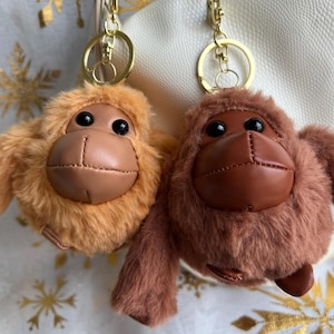 May include: Two plush orangutan keychains. One is light brown, the other is dark brown. Both have leather-like faces and black eyes. The keychains have gold-colored metal rings.