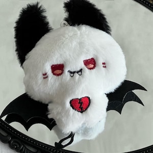 May include: A plush toy shaped like a skull with white fur, black fuzzy ears, and black bat wings. The face features embroidered red eyes and small fangs. A red broken heart is stitched onto the center of the skull, and a small black tail hangs below.