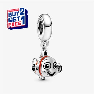 Puede incluir: Un charm de plata con un pez de dibujos animados sonriente con rayas naranjas y blancas. El charm cuelga de un lazo plateado con el nombre de la marca. Un gráfico rojo, blanco y azul con el texto "BUY 2 GET 1 FREE" está en la esquina superior izquierda.
