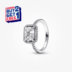 Puede incluir: Un anillo plateado con una gema rectangular y transparente en el centro, rodeada de piedras claras más pequeñas. La banda también está adornada con pequeñas piedras claras. El anillo tiene la inscripción "PANDORA". Un banner promocional "BUY 2 GET 1 FREE" está en la parte superior izquierda.