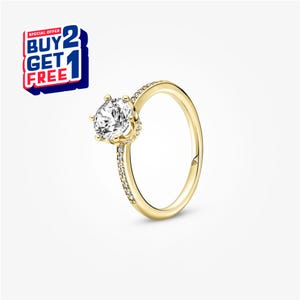 May include: A gold-colored ring with a large, clear, round gemstone at the center. The band is also gold and has small, clear gemstones set along one side. The image includes a "BUY 2 GET 1 FREE" promotional offer.