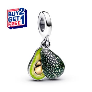 Può includere: Un charm in argento con due metà di avocado. Una metà è verde con un nocciolo dorato, l'altra è verde scuro con puntini bianchi. Il charm ha un anello d'argento per attaccarlo a un braccialetto o una collana. Un'offerta "COMPRA 2 PRENDI 1 GRATIS" è nell'angolo in alto a sinistra.