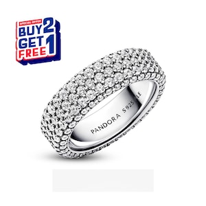 Puede incluir: Un anillo plateado con tres filas de piedras claras y brillantes. El anillo está grabado con "PANDORA S925 ALE". Una oferta de "COMPRA 2 Y LLÉVATE 1 GRATIS" se muestra en la esquina superior izquierda.