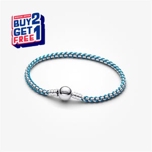 Op de afbeelding: Een zilveren armband met een geweven blauw koordontwerp. De armband heeft een ronde sluiting met de merknaam. Een speciale aanbieding grafiek met de tekst "BUY 2 GET 1 FREE" staat in de linkerbovenhoek.