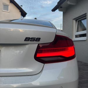 K&ouml;nnte beinhalten: Die R&uuml;ckansicht eines wei&szlig;en Autos mit einem schwarzen B58-Emblem auf dem Kofferraumdeckel. Das Fahrzeug verf&uuml;gt &uuml;ber eine rote R&uuml;ckleuchteneinheit mit einem markanten horizontalen Lichtbalken und einen kleinen runden Parksensor am Sto&szlig;f&auml;nger.
