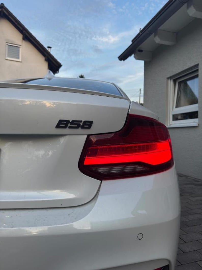 K&ouml;nnte beinhalten: Die R&uuml;ckansicht eines wei&szlig;en Autos mit einem schwarzen B58-Emblem auf dem Kofferraumdeckel. Das Fahrzeug verf&uuml;gt &uuml;ber eine rote R&uuml;ckleuchteneinheit mit einem markanten horizontalen Lichtbalken und einen kleinen runden Parksensor am Sto&szlig;f&auml;nger.