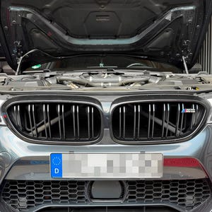 Puede incluir: Un sedán BMW M5 de color gris oscuro con el capó abierto, dejando ver el compartimento del motor. El frontal presenta una parrilla negra de doble riñón con una insignia M, una toma de aire inferior de panal y una matrícula alemana.