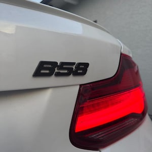K&ouml;nnte beinhalten: Ein wei&szlig;er Kofferraumdeckel mit einem schwarzen B58-Emblem, das &uuml;ber einer leuchtend roten R&uuml;ckleuchte angebracht ist. Die Oberfl&auml;che ist glatt und gl&auml;nzend und zeigt die hintere Ecke des Fahrzeugs mit einer dezenten Spoilerkante am oberen Rand.