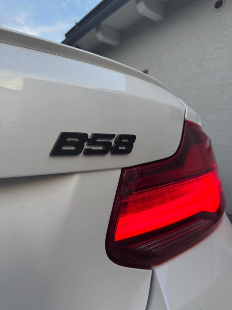 K&ouml;nnte beinhalten: Ein wei&szlig;er Kofferraumdeckel mit einem schwarzen B58-Emblem, das &uuml;ber einer leuchtend roten R&uuml;ckleuchte angebracht ist. Die Oberfl&auml;che ist glatt und gl&auml;nzend und zeigt die hintere Ecke des Fahrzeugs mit einer dezenten Spoilerkante am oberen Rand.